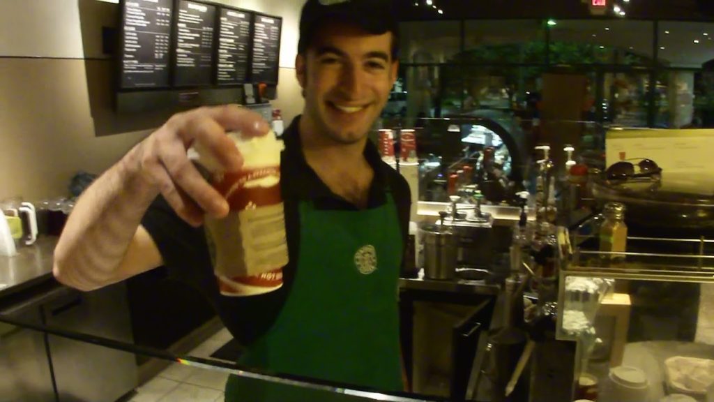 Bryan Kofsky, in Starbucks uniform with a green apron and black hat, smiling and handing over a holiday Starbucks coffee cup from behind the counter with an espresso machine in the background, in 2010.