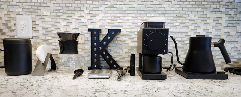 Coffee setup with Fellow and Hario equipment in matte black on a kitchen counter with glass tile backsplash, taken in June 2023.